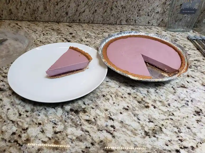 A slice of purple pie sits on a white plate next to the aluminum pie tin, which still contains the majority of the pie. The…