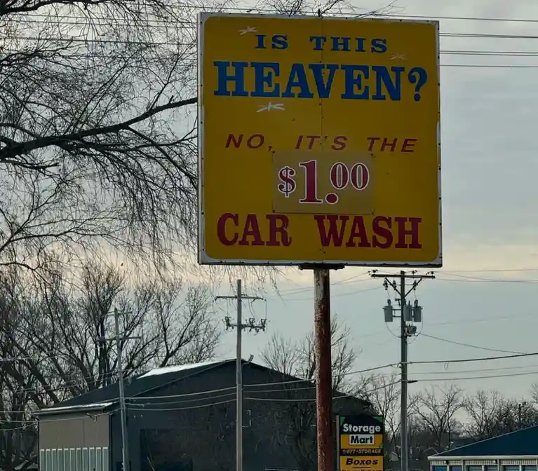 A yellow roadside sign with blue and red text asks if the location is heaven, then humorously answers it's a $1.00 car wash…
