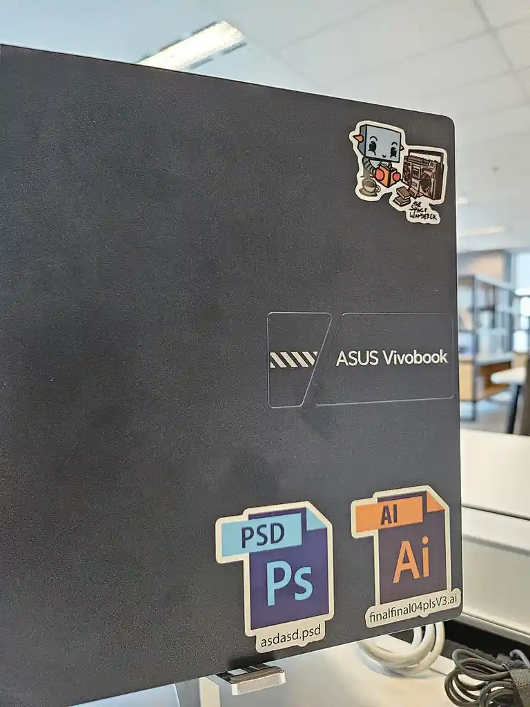 A close-up of a black ASUS Vivobook laptop lid decorated with stickers. Two prominent stickers mimic Adobe Photoshop (PSD)…