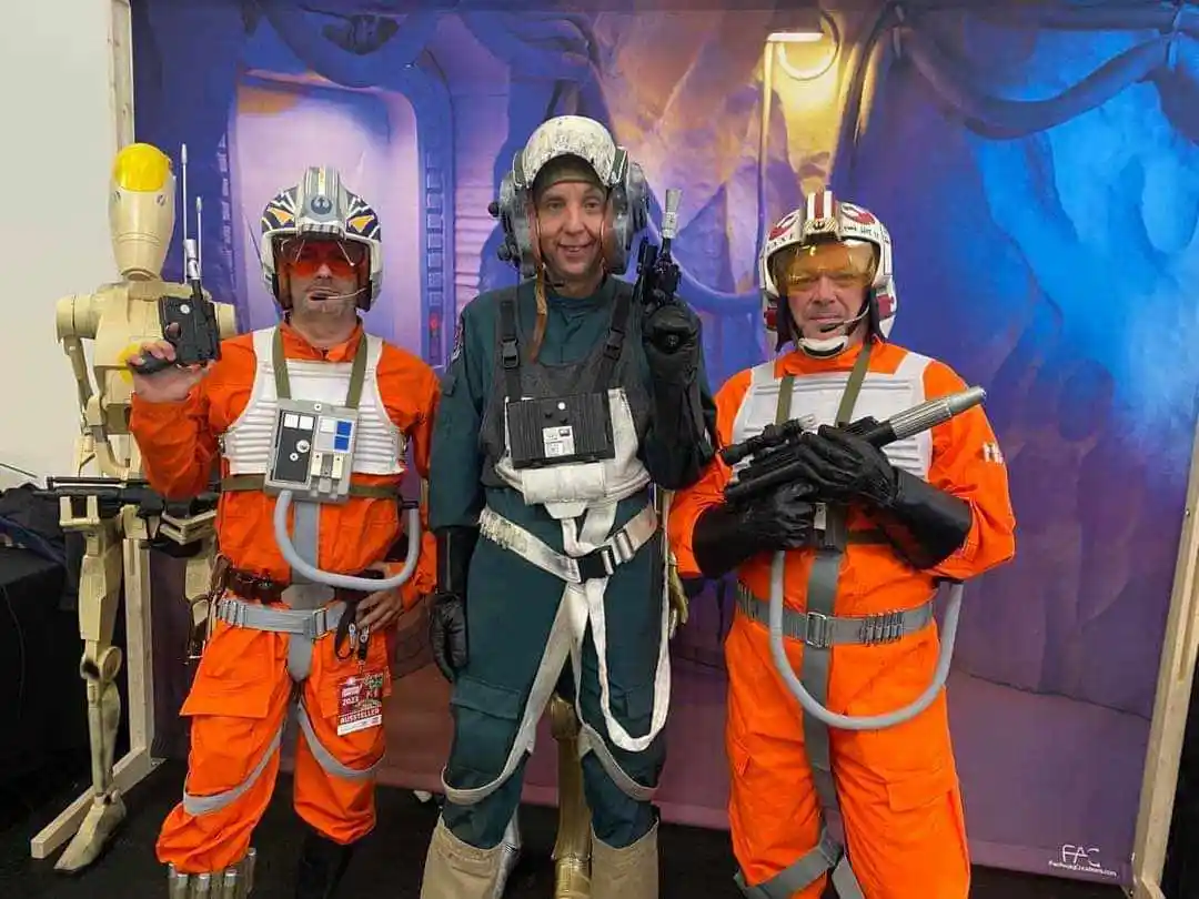 Three men dressed as Star Wars pilots, one in green and two in orange flight suits, pose with props and a droid. The man in…
