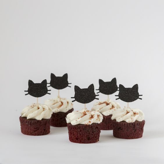 Five red velvet cupcakes with white frosting and brown sprinkles are arranged on a plain white surface, each topped with a…