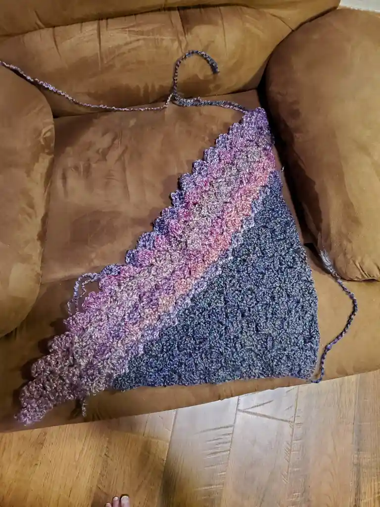 A partially completed, triangular crochet blanket with a gradient of purple, pink, and blue yarn rests on a brown couch. The…