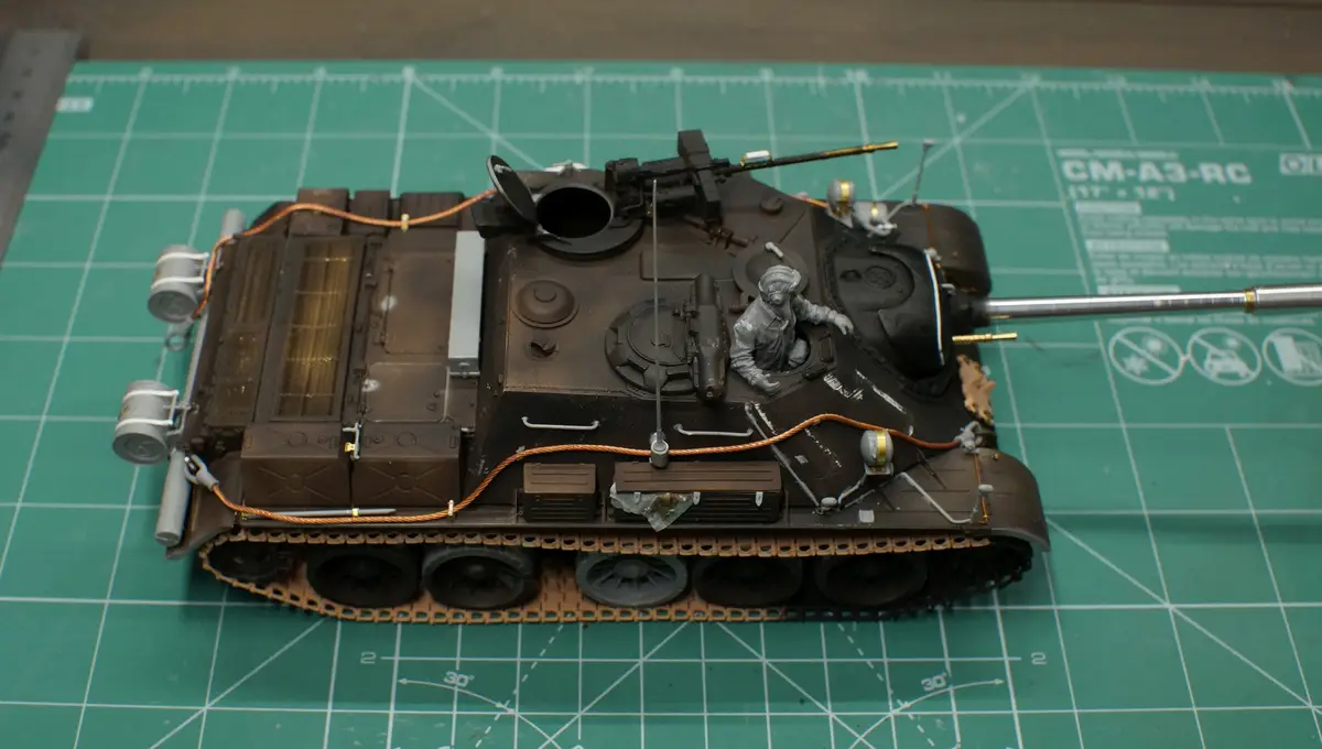 A detailed scale model of a military tank sits on a green cutting mat with grid lines and measurements. The tank is dark…