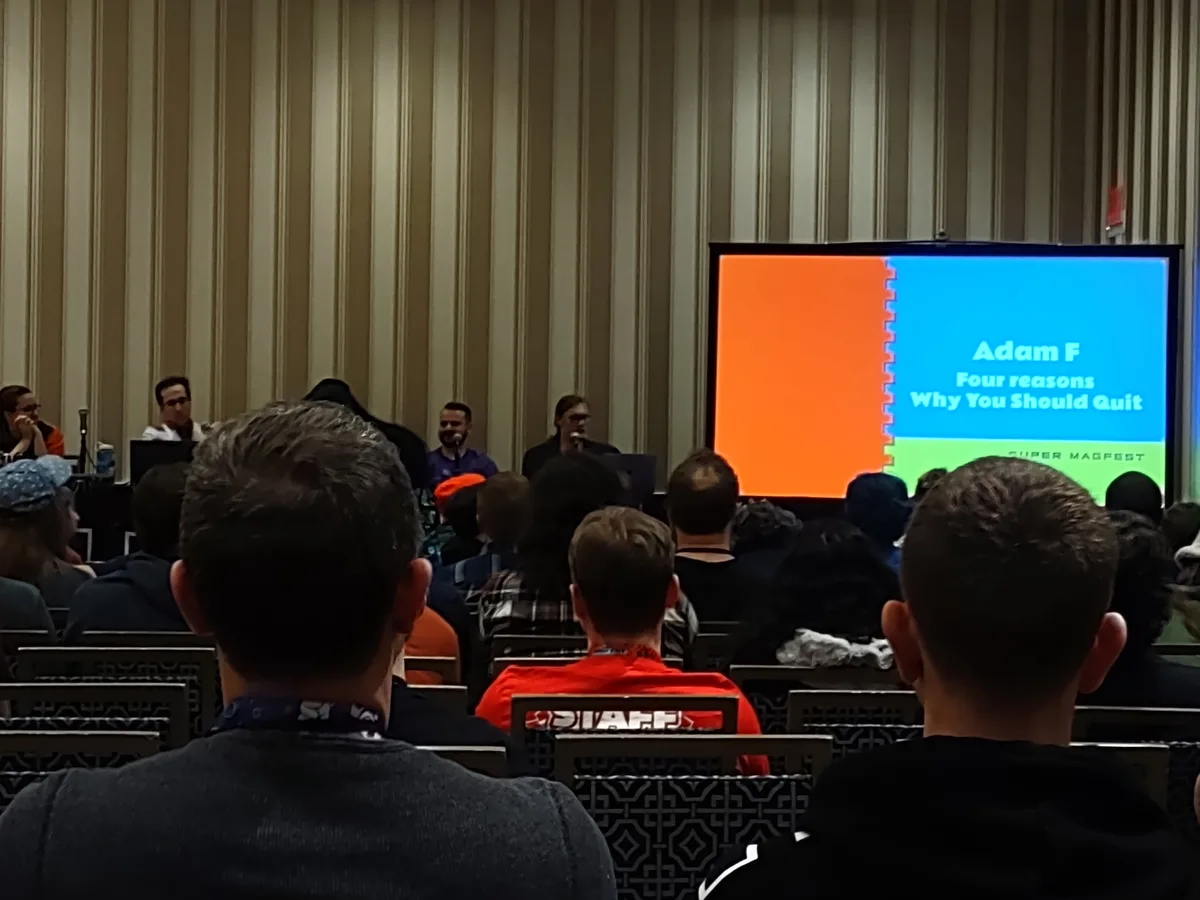 An audience watches a presentation at a conference, with a large screen displaying a slide titled 'Adam F Four reasons Why…