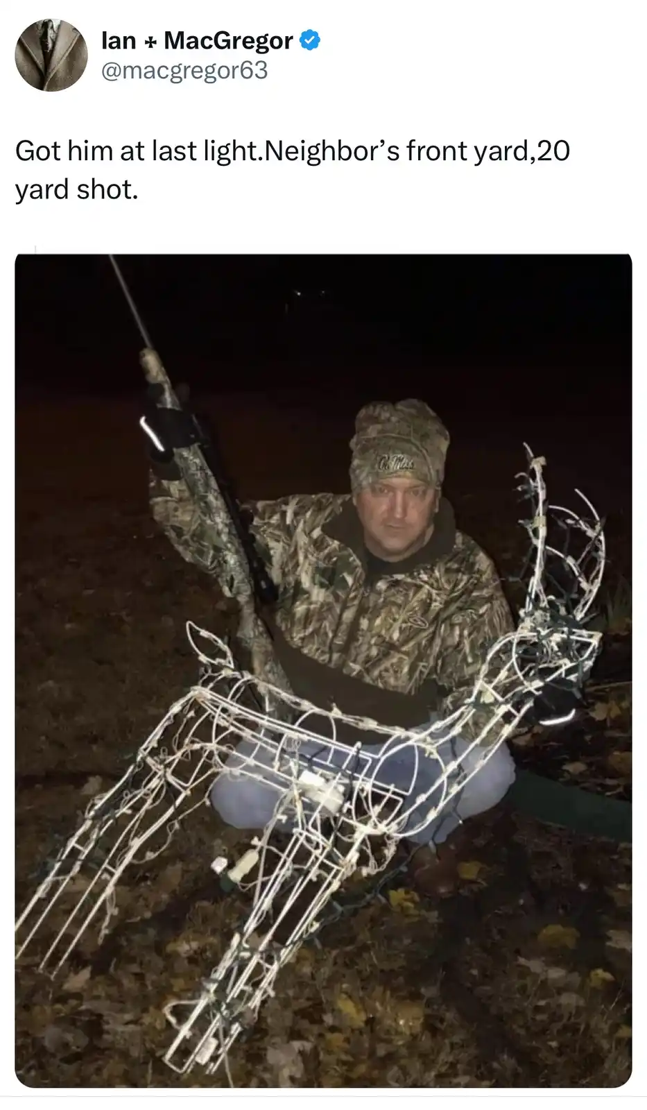 A man in camouflage clothing and a hat kneels outdoors at night, holding a rifle while posing beside a wireframe deer…