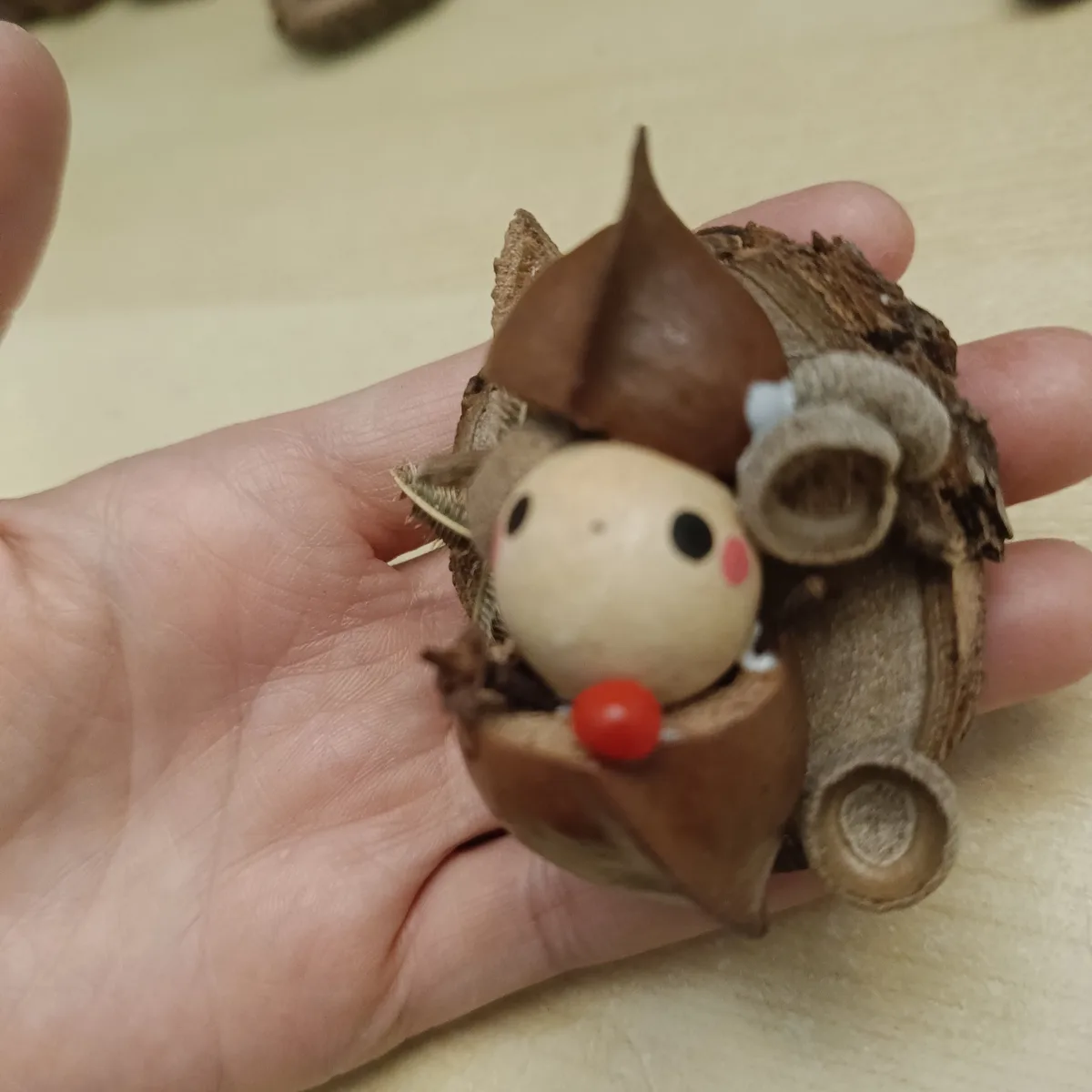 A hand holds a small, handmade figurine resembling a forest gnome, crafted from natural materials like acorn caps and dried…