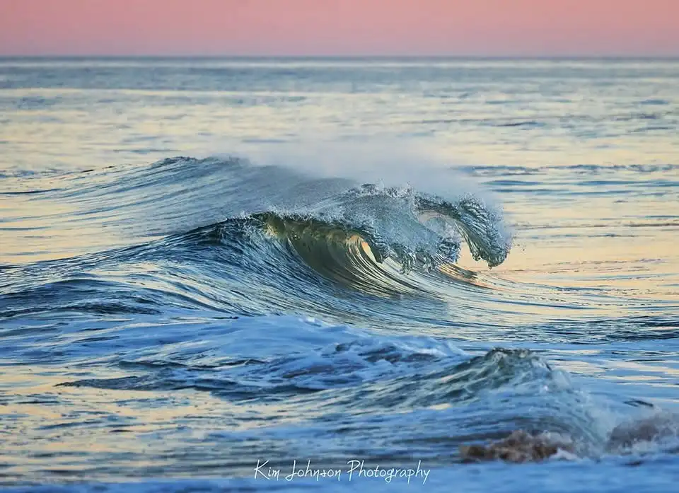 A serene ocean wave curls gracefully under a soft pink and orange sunset sky, with the photographer's watermark 'Kim Johnson…