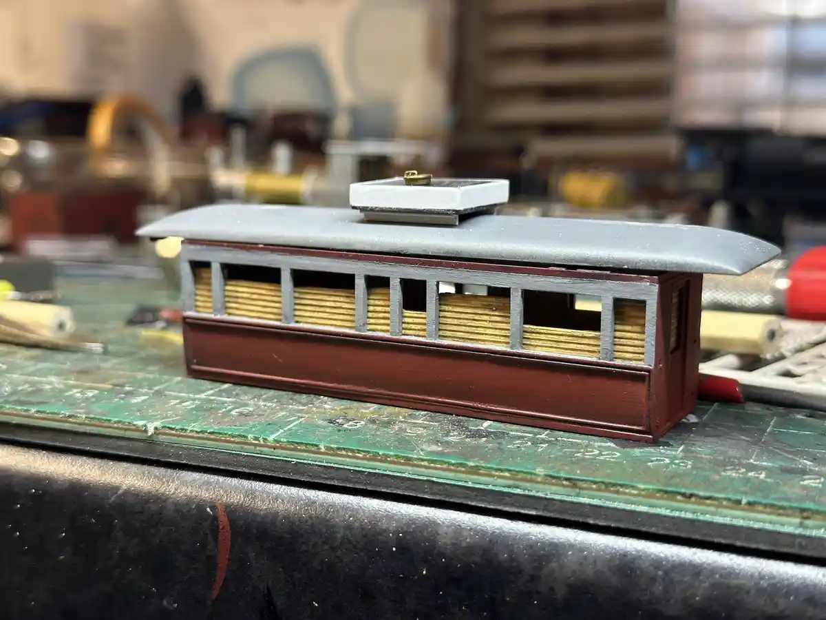 A close-up photo shows a small, partially painted model trolley car with a grey roof and maroon body, sitting on a green…