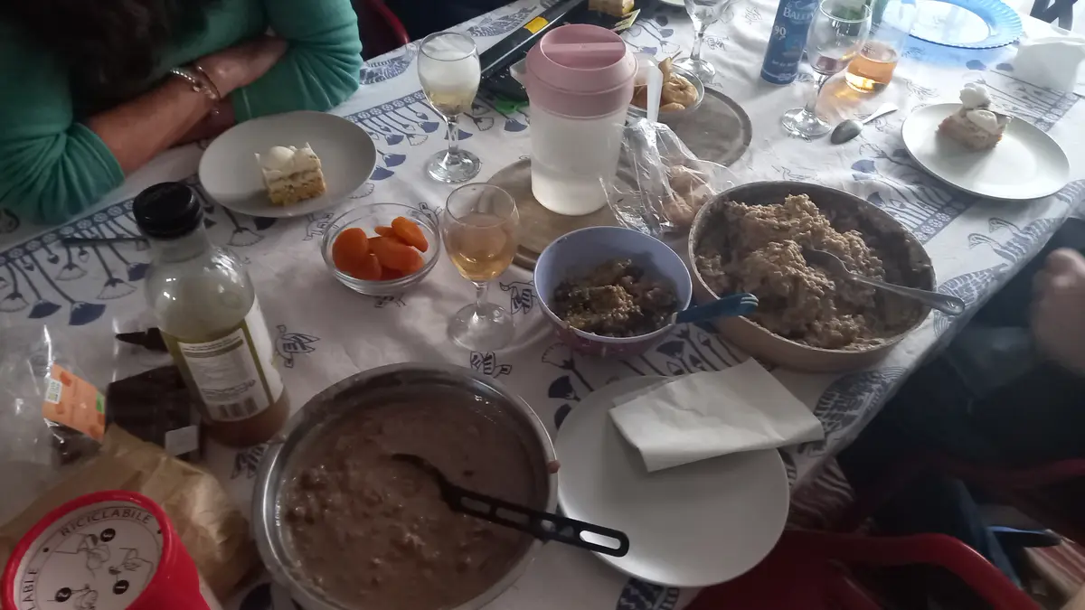 An overhead shot of a cluttered dining table with bowls of oatmeal, cake slices, wine glasses, and various snacks. Two…