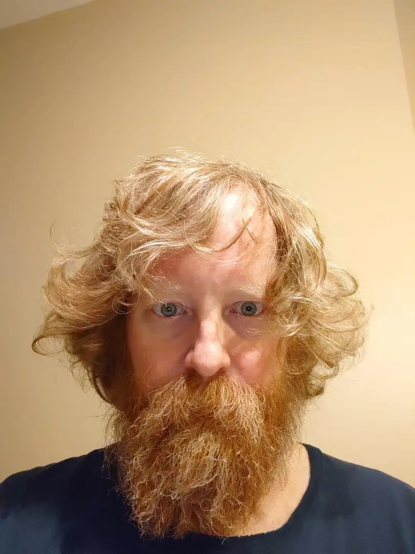 A man with long, wavy blonde hair and a full, reddish-brown beard stares directly at the camera with wide, slightly startled…