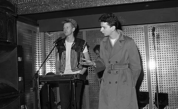 A black-and-white photo shows two men on a stage; one in a striped vest plays a keyboard while the other in a trench coat…