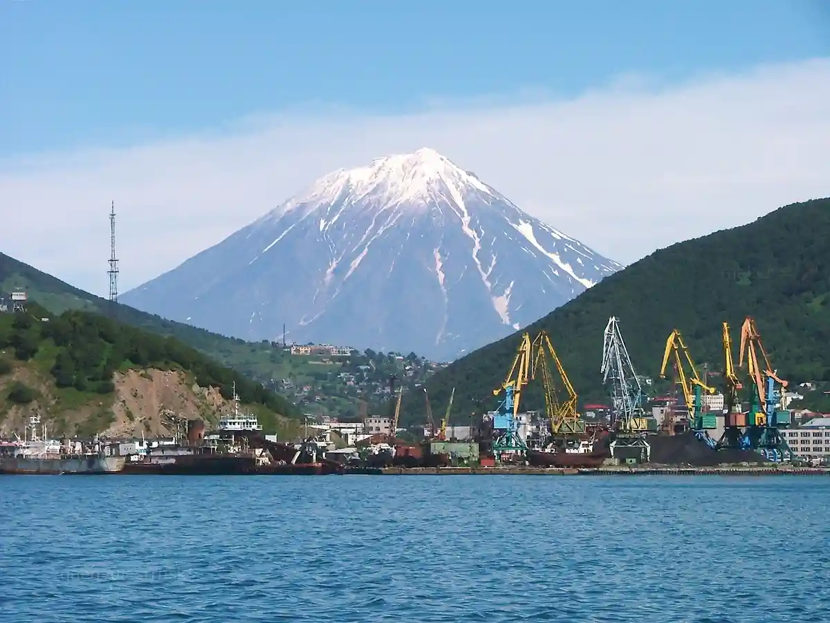 A scenic view of a snow-capped mountain rising behind a harbor with industrial cranes and ships. The overlaid text reads…