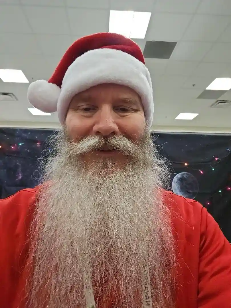 A man with a long, gray beard and mustache wears a red and white Santa hat and a red shirt, looking at the camera with a…