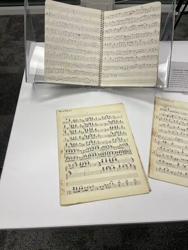 The image shows three handwritten musical scores on paper, displayed on a white table. One score is in a clear acrylic…