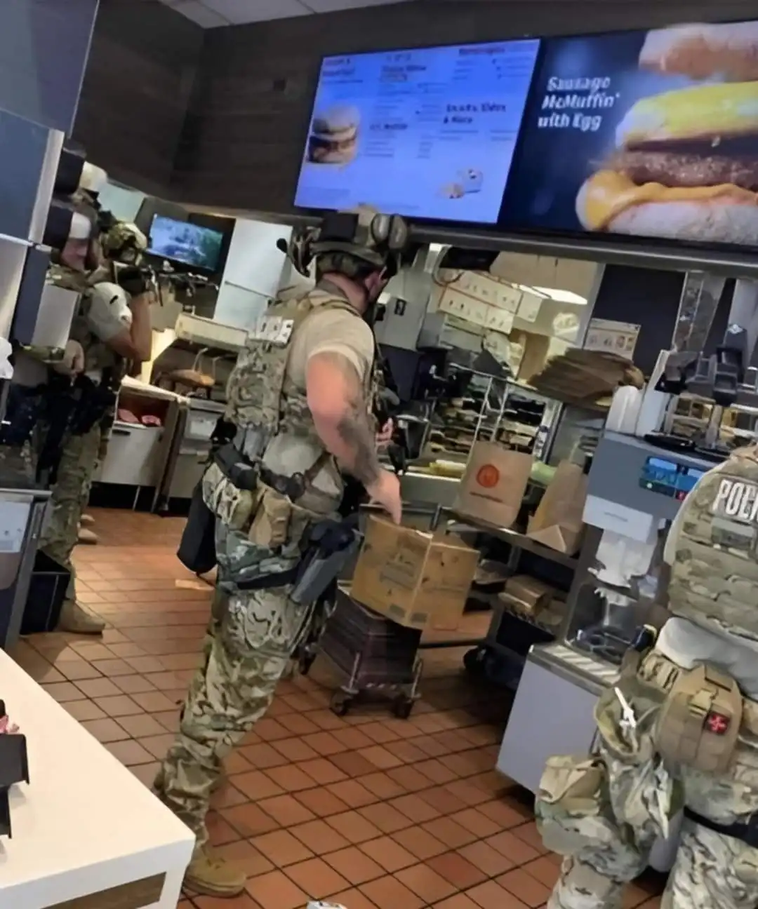 Three armed police officers in tactical gear are inside a McDonald's restaurant, one pushing a cart with a cardboard box…