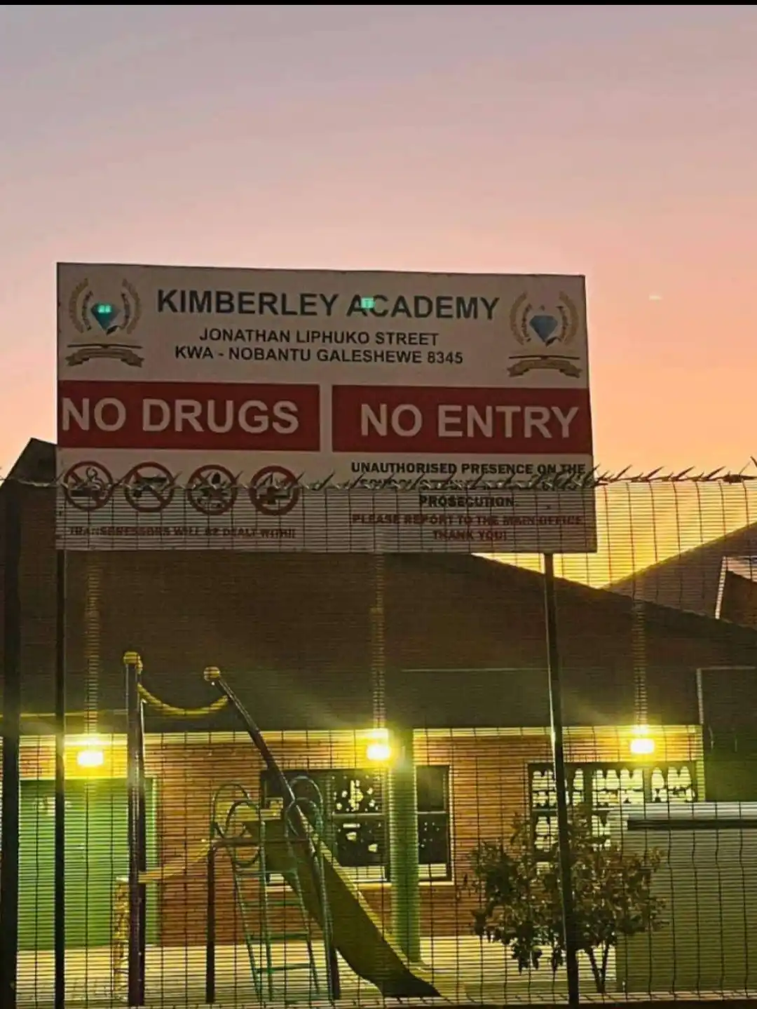 A large sign for Kimberley Academy at dusk shows 'NO DRUGS' and 'NO ENTRY' in bold red boxes. Below, smaller text warns of…