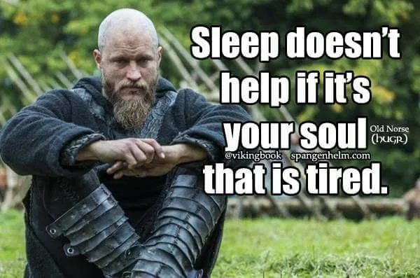 A bald, bearded Viking warrior in dark clothing and leather armor sits on grass with a serious expression, hands clasped…
