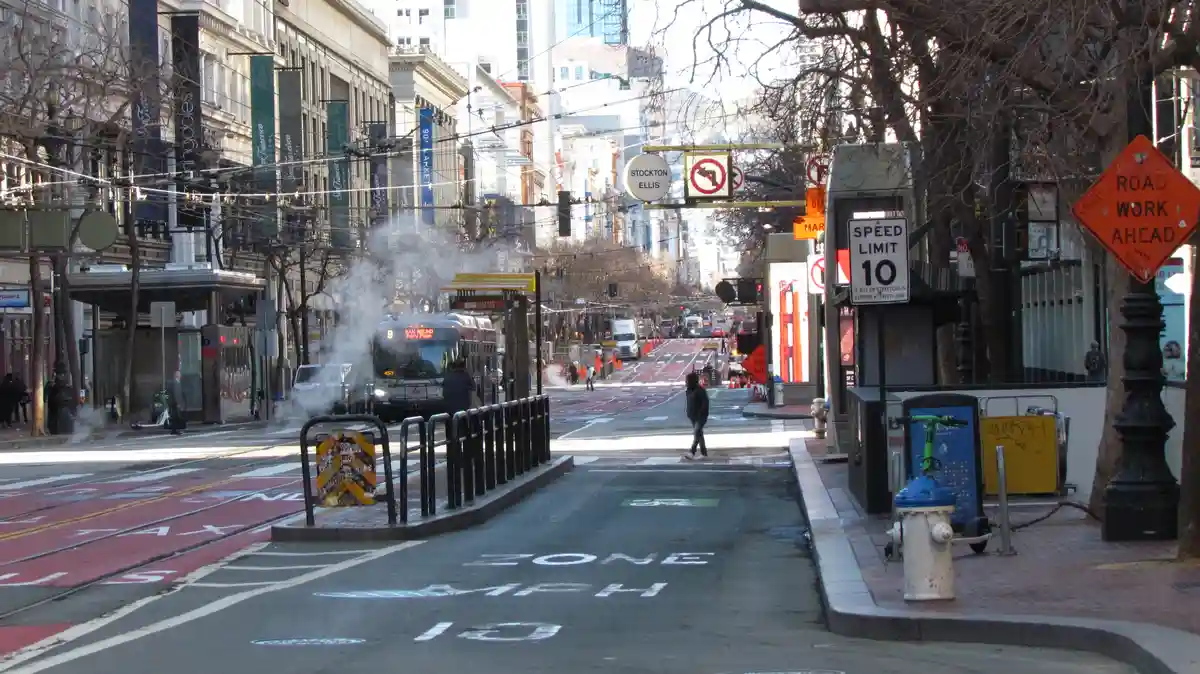 A street scene in San Francisco with a bus emitting steam, a "SPEED LIMIT 10" sign, and "ZONE 10 MPH" painted on the road…