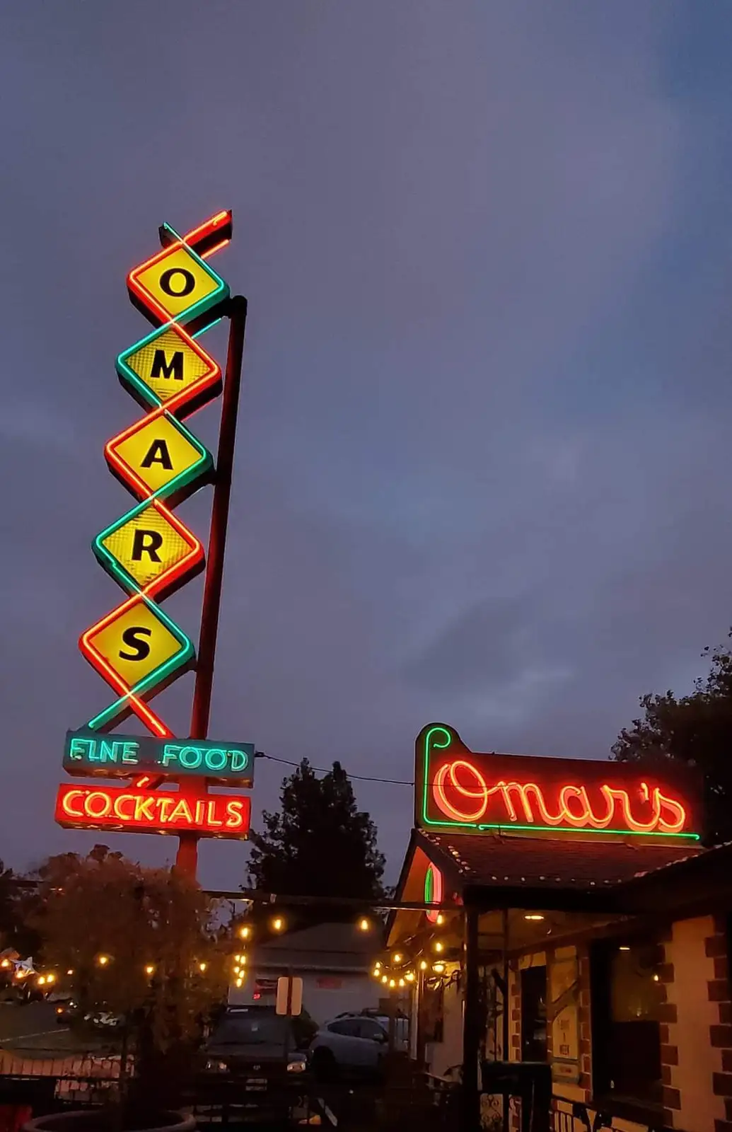 A tall, vertical neon sign spells out 'OMAR'S' in diamond-shaped segments with red, green, and yellow lighting against a…