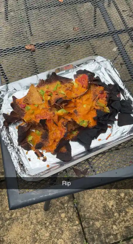 A tray of nachos covered in melted cheese and toppings sits on a metal mesh table, with many tortilla chips visibly burnt…