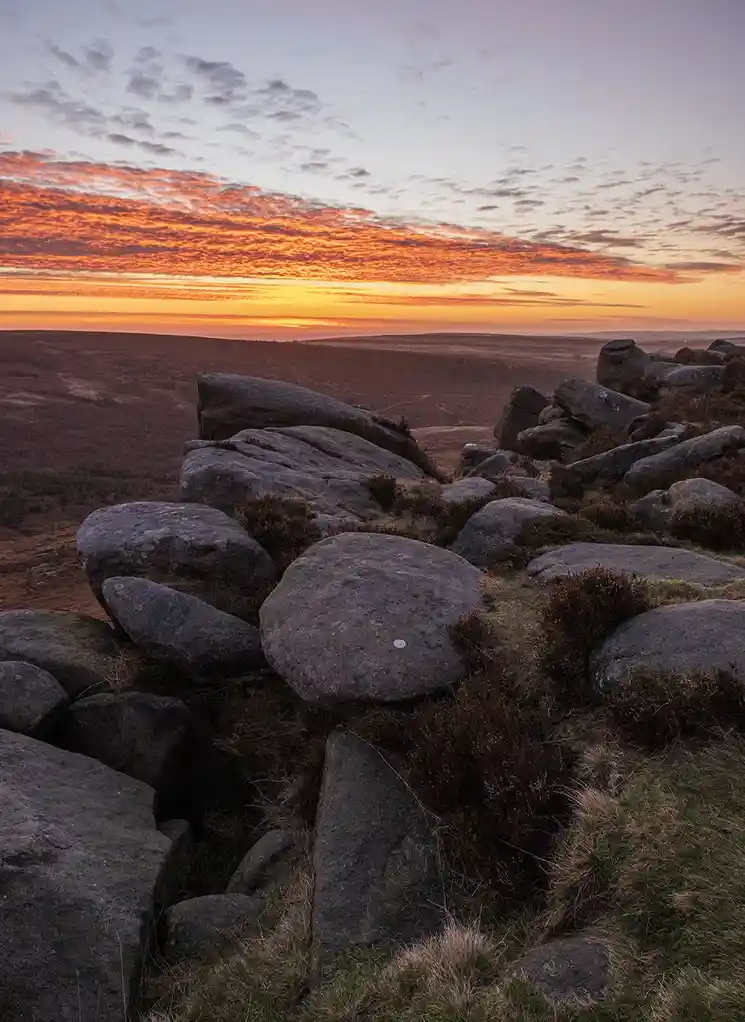 A scenic photo of a rocky hilltop at sunrise or sunset, with vibrant orange and pink clouds above a vast, rolling landscape…