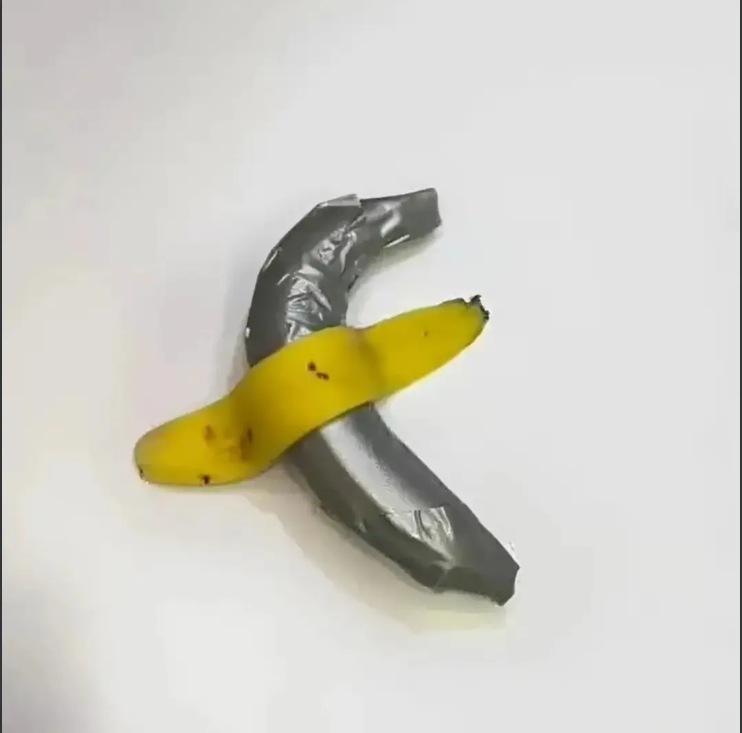 A single yellow banana with brown spots is taped crosswise to another banana that's been wrapped entirely in gray duct tape…