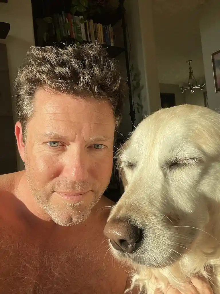 A shirtless man with salt-and-pepper hair and blue eyes looks at the camera with a slight smirk, while a golden retriever…