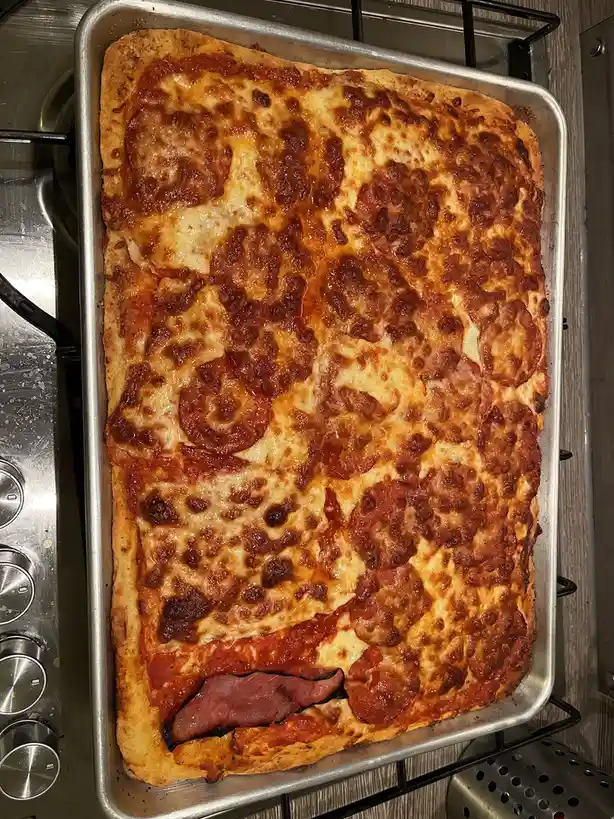 A rectangular pepperoni pizza on a metal baking sheet sits on a stovetop, with melted cheese, sauce, and pepperoni. The…