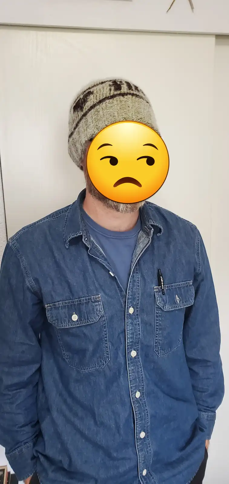 A person wearing a beige and brown striped knit beanie, a blue denim shirt over a blue t-shirt, and a black pen in their…