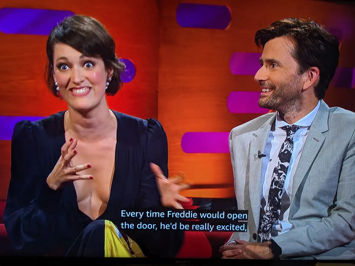 A woman with short dark hair and a dark blue dress gestures animatedly while speaking, her hand over her chest, as a man in…