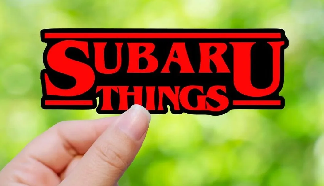 A hand holds up a red and black sticker with the text 'SUBARU THINGS' in a bold, stylized font reminiscent of the 'Stranger…