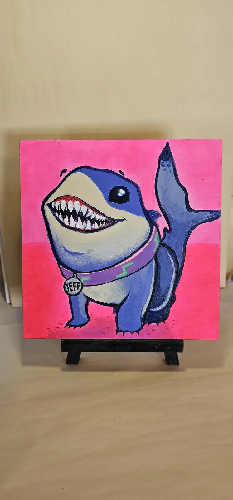 A hand-painted cartoon shark with a wide, toothy grin and a purple collar stands on a black easel against a bright pink…