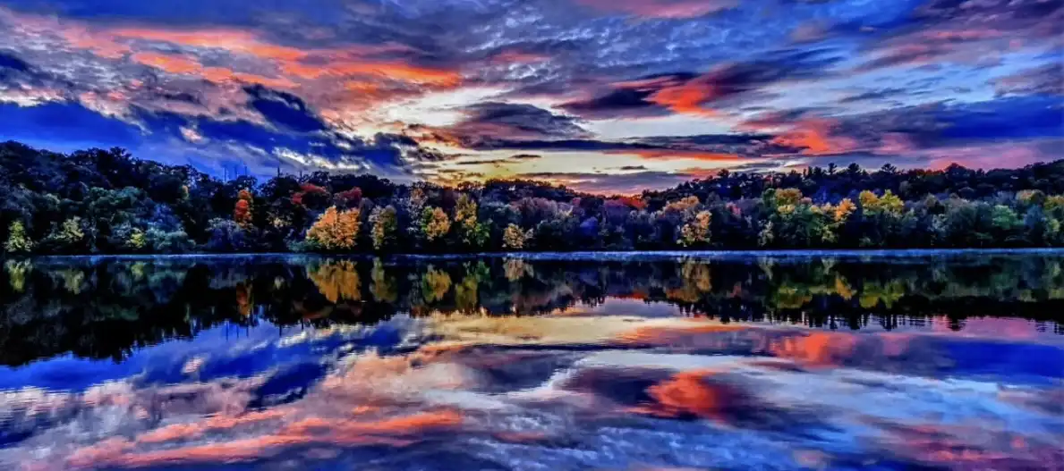 A vibrant, highly saturated landscape photo shows a lake reflecting a dramatic sunset sky filled with orange, pink, and deep…