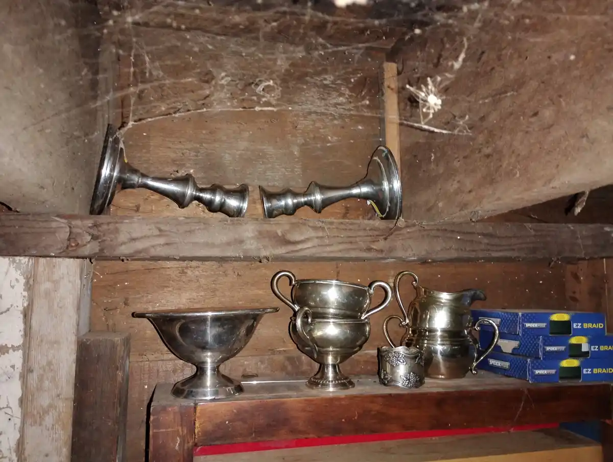 A dusty attic shelf holds tarnished silverware including candlesticks, a bowl, a sugar bowl, a creamer, and a small cup…