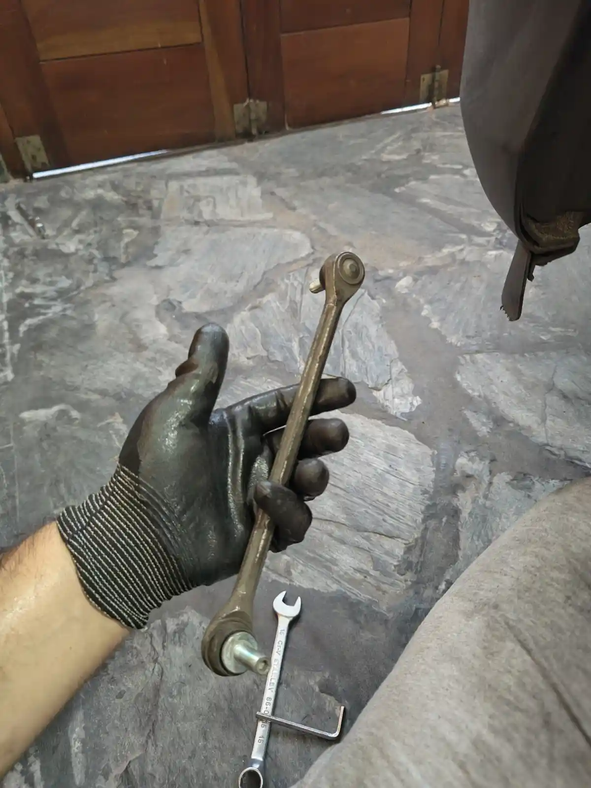 A gloved hand holds a wrench covered in grease, with another wrench lying on a stone floor near a car part. The overlaid…