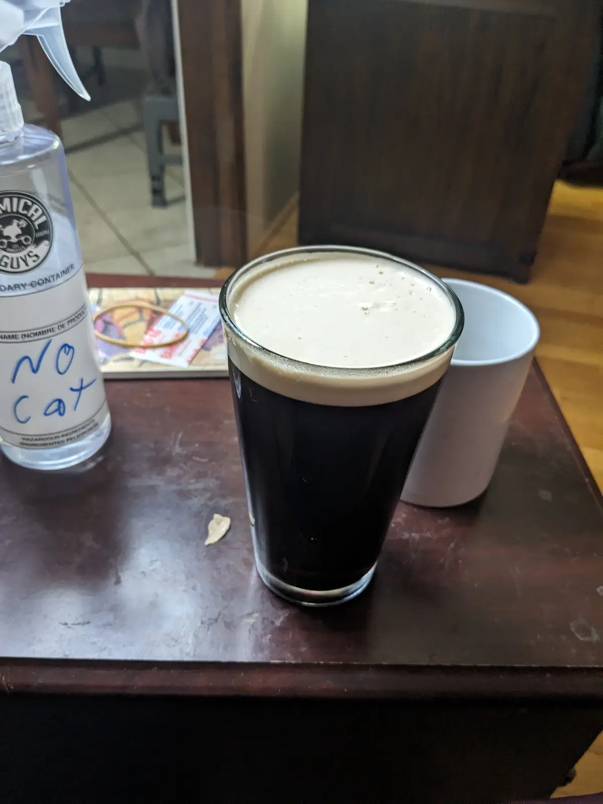 A glass of dark beer with a thick creamy head sits on a wooden table next to a white mug and a spray bottle labeled…