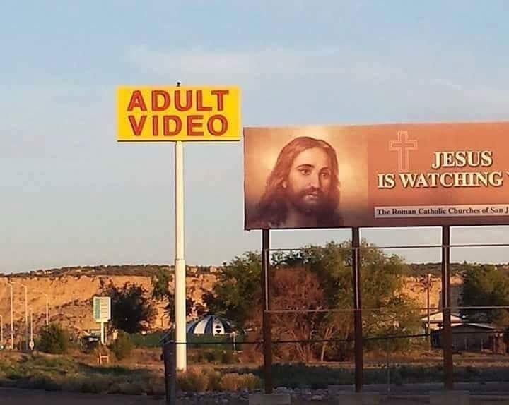 Two large roadside signs stand side by side: one is a yellow sign with red text reading 'ADULT VIDEO', and the other is a…