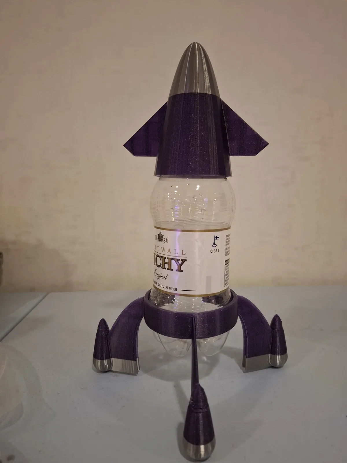 A clear plastic bottle of Vichy water is modified into a rocket with purple 3D-printed parts, including a pointed nose cone…