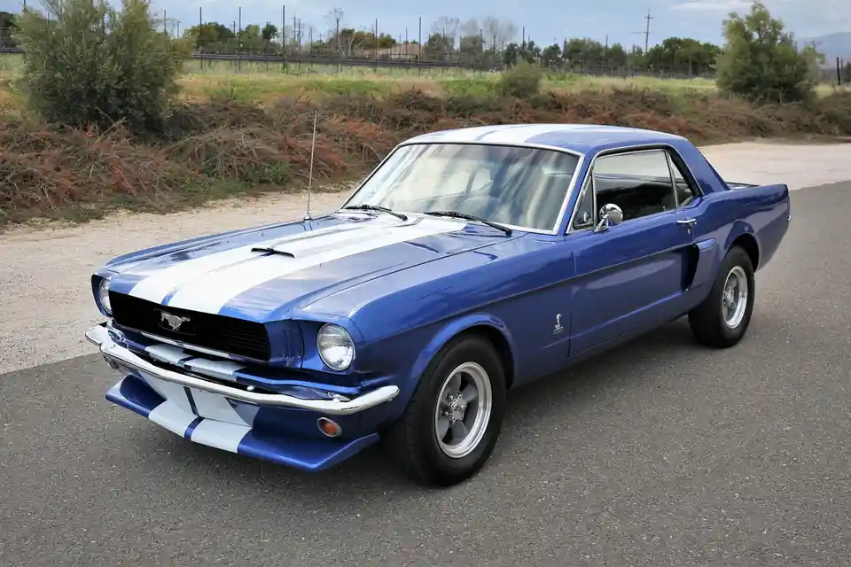 A blue 1960s Ford Mustang Shelby GT350 with white racing stripes is parked on an asphalt road, featuring chrome bumpers…