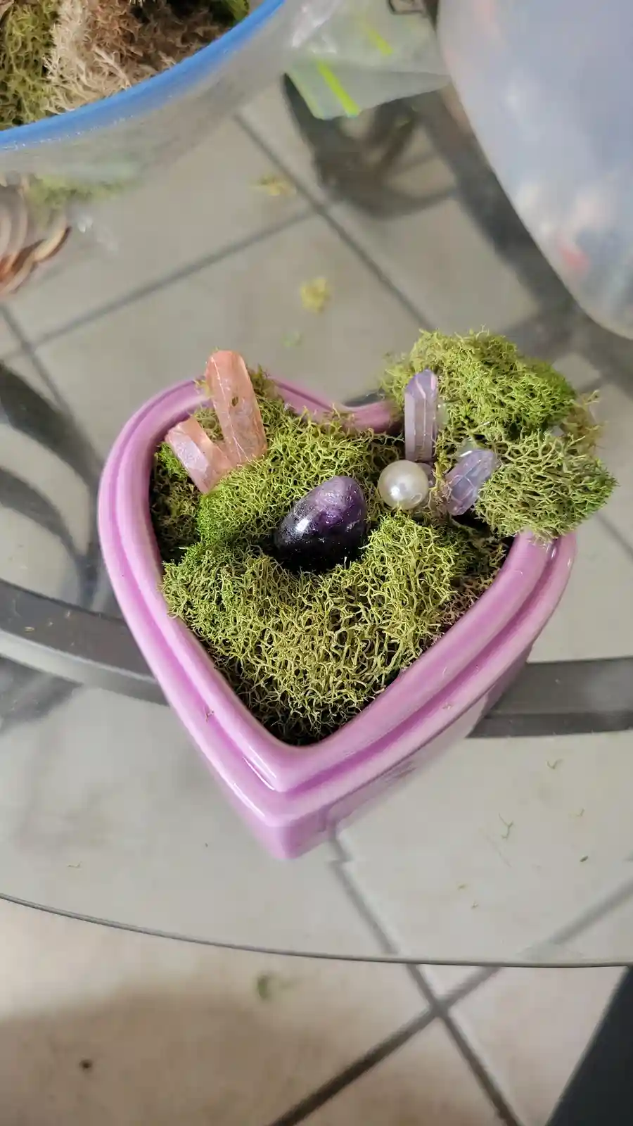A pink ceramic heart-shaped planter filled with green moss, featuring a central purple crystal, a pearl, and several smaller…