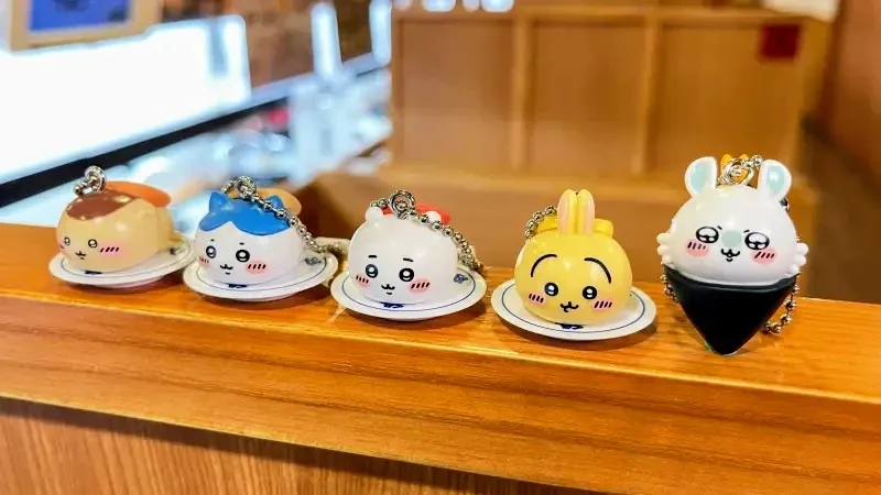 Five small, cute, cartoonish keychains shaped like Japanese food items (onigiri, tamagoyaki, rice ball, egg, and onigiri…