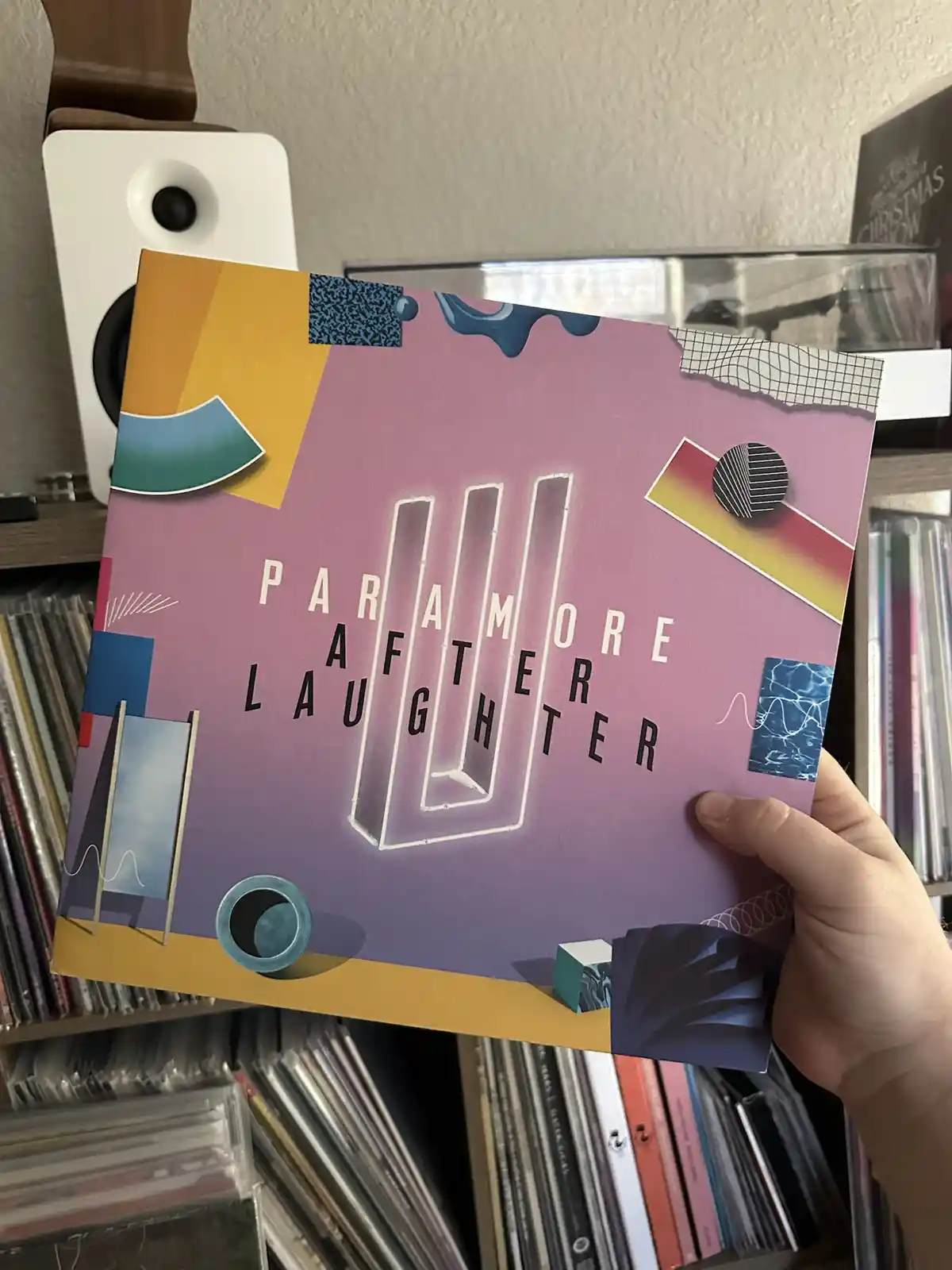 A hand holds up the album cover for Paramore's 'After Laughter', which features a vibrant, abstract design with geometric…