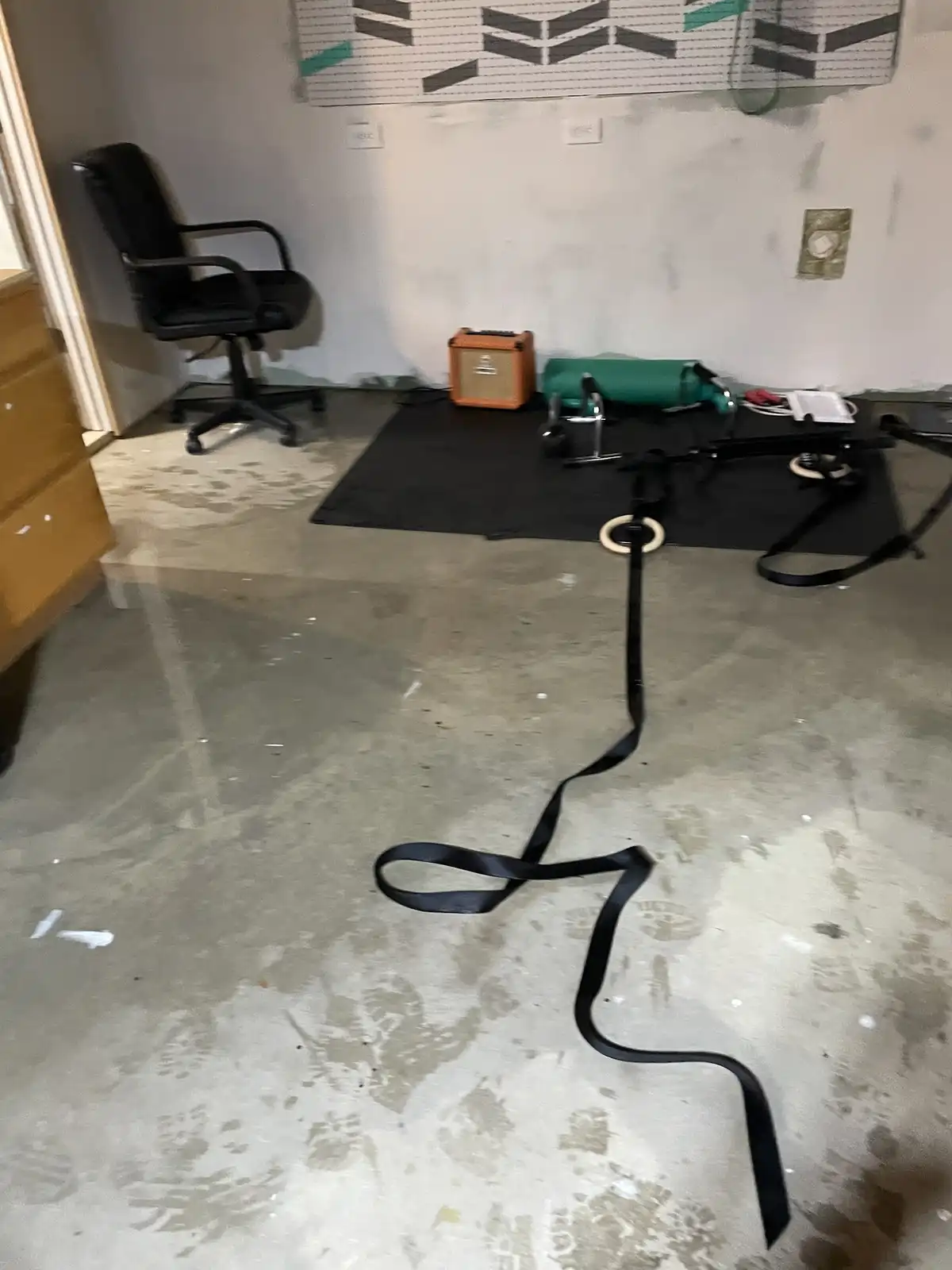 A messy room with a concrete floor covered in footprints and scattered debris. A black office chair sits in the corner…