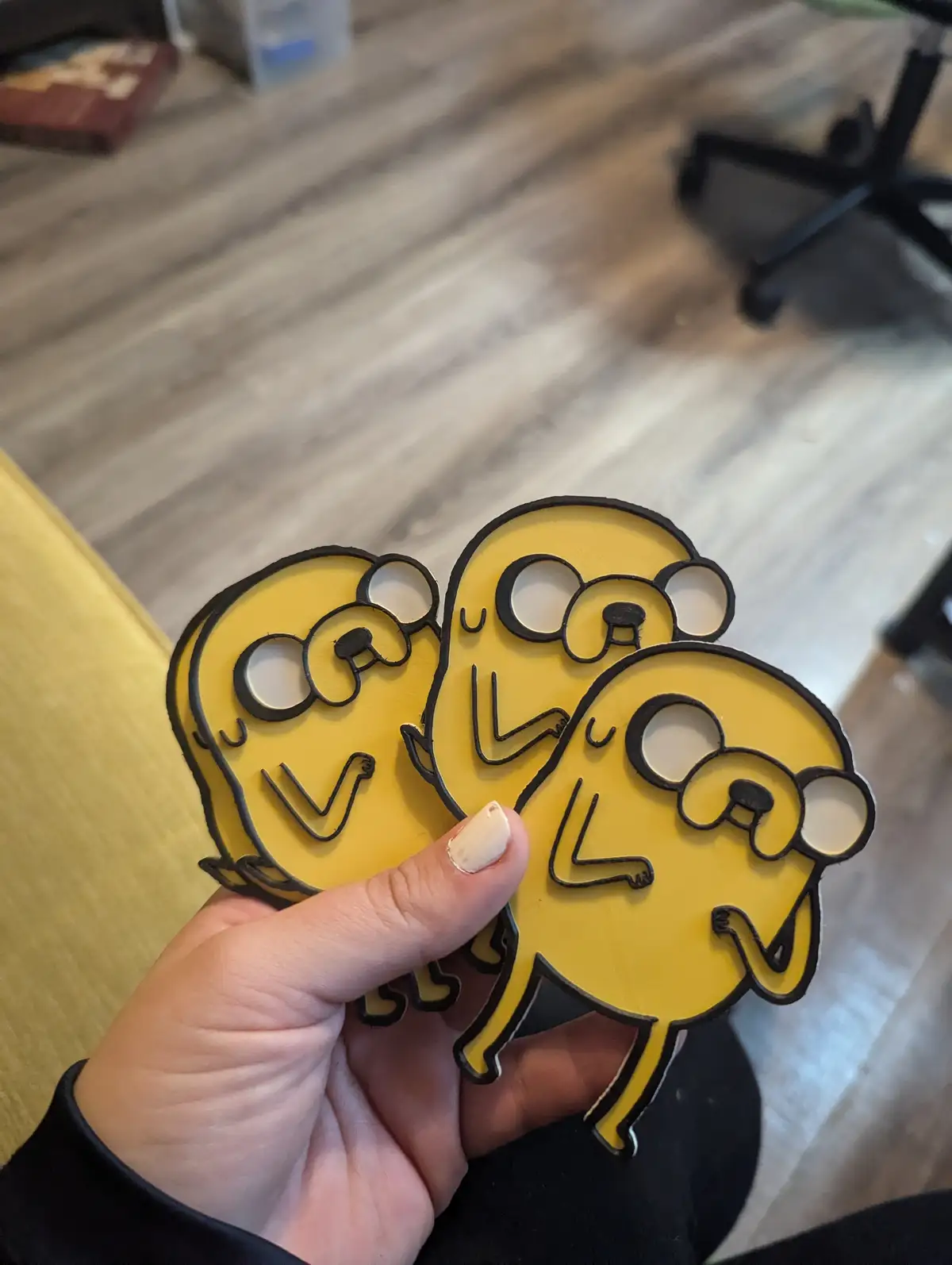 A hand holds three identical yellow cardboard cutouts of the cartoon character Jake from Adventure Time, each with a simple…