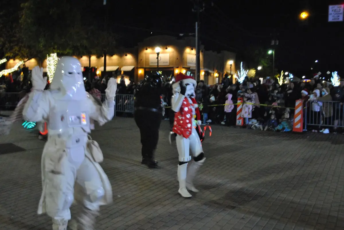 At a nighttime holiday parade, two people in Star Wars Stormtrooper costumes walk among spectators. One trooper wears a red…
