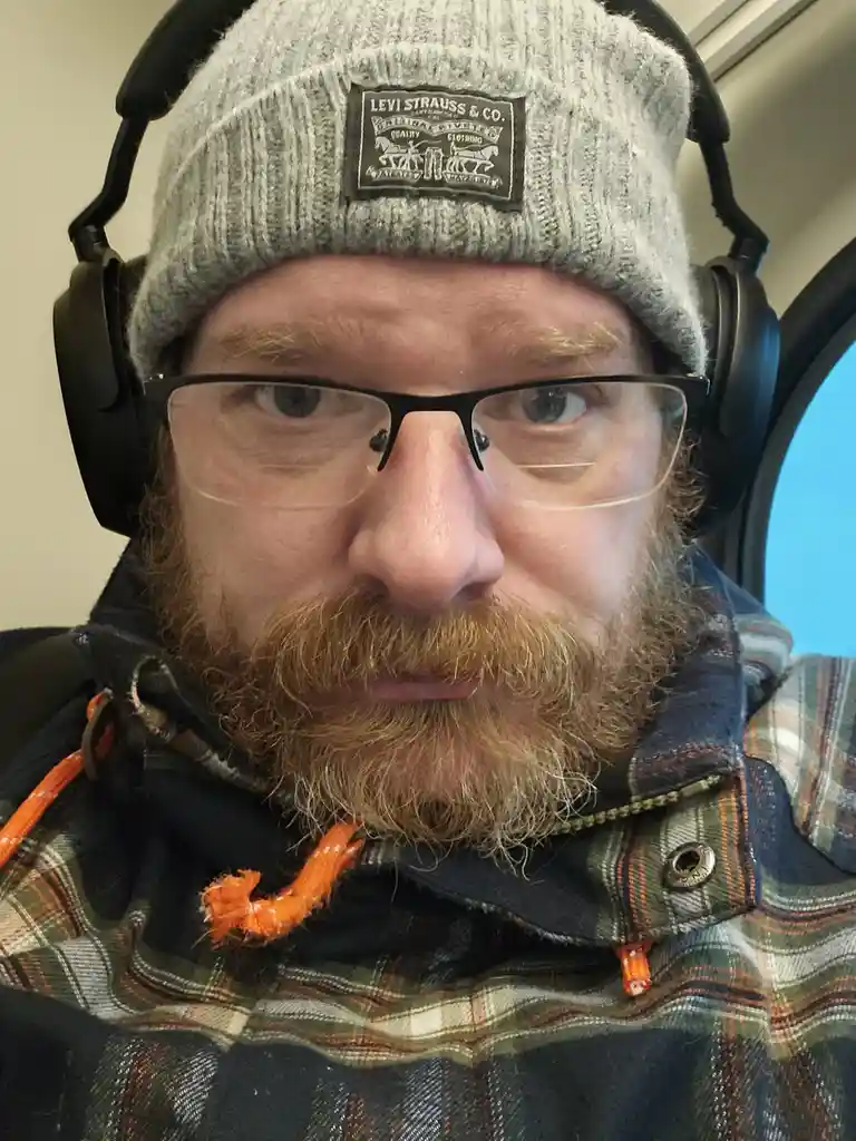 A man with a reddish beard and glasses wears a grey Levi's beanie, black over-ear headphones, and a plaid jacket with orange…