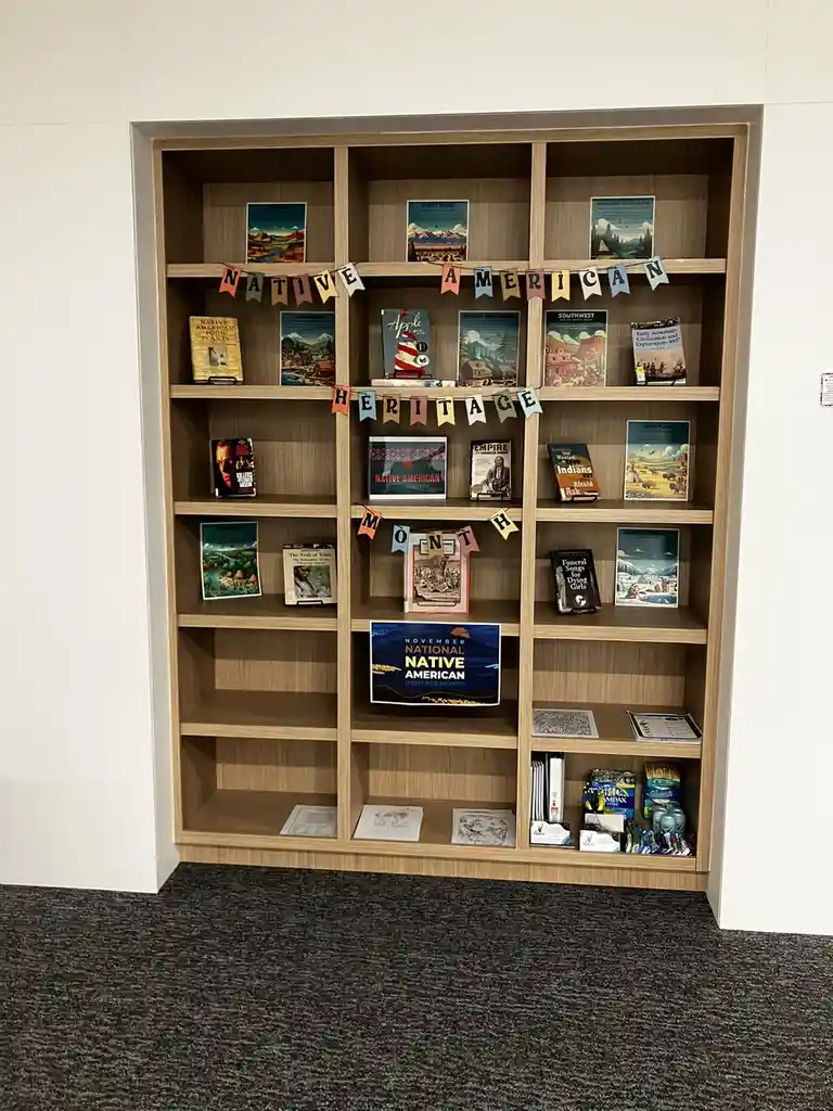 A wooden bookshelf displays books and posters themed around Native American heritage, with colorful banners spelling 'NATIVE…