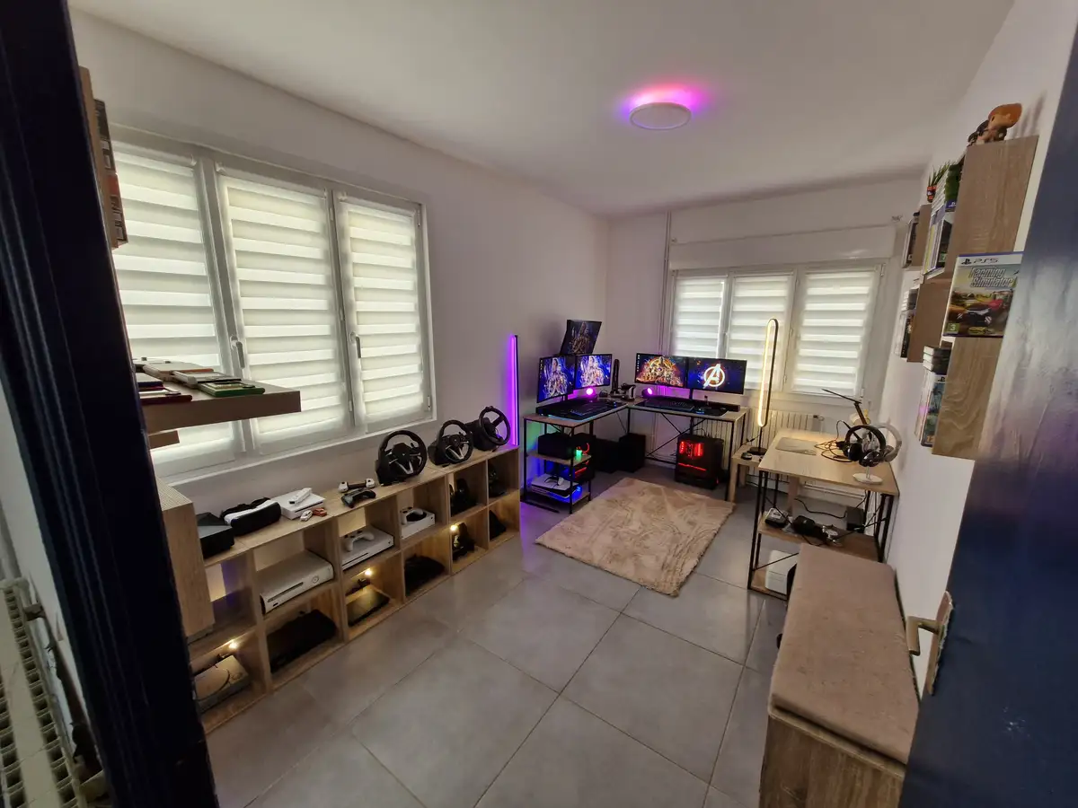 A well-lit, modern gaming room with multiple setups: a central desk with three monitors displaying colorful game scenes, a…