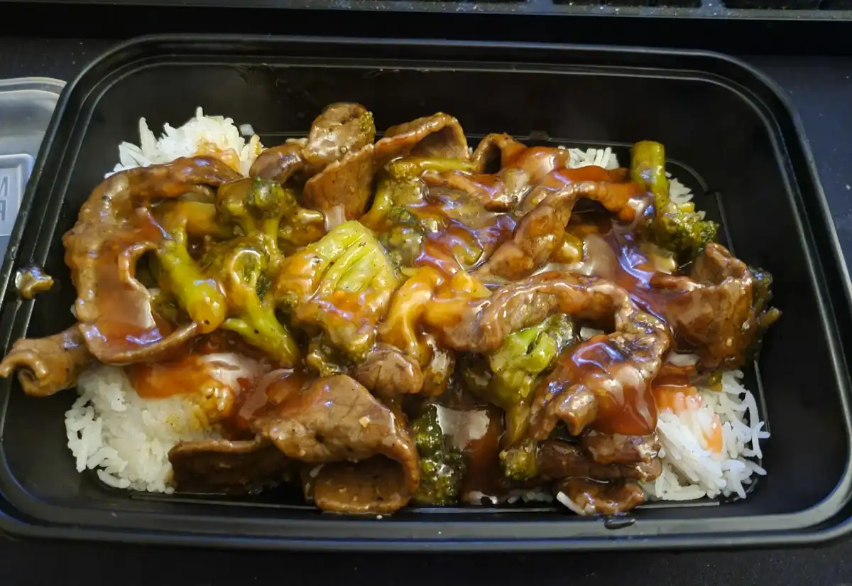A close-up photo of a black takeout container filled with beef and broccoli stir-fry generously covered in sauce, served…