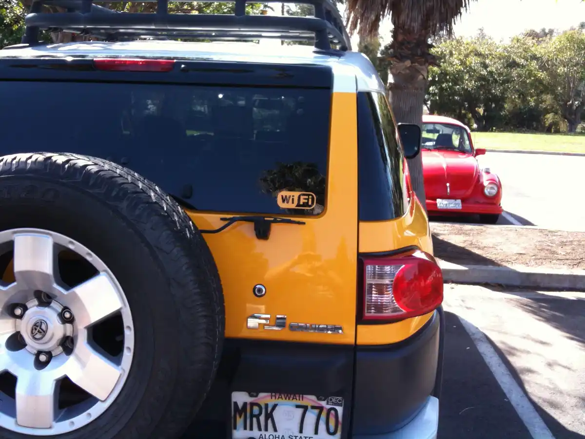 The image shows the rear of a yellow and black Toyota FJ Cruiser with a spare tire mounted on the back, a 'wifi' sticker on…